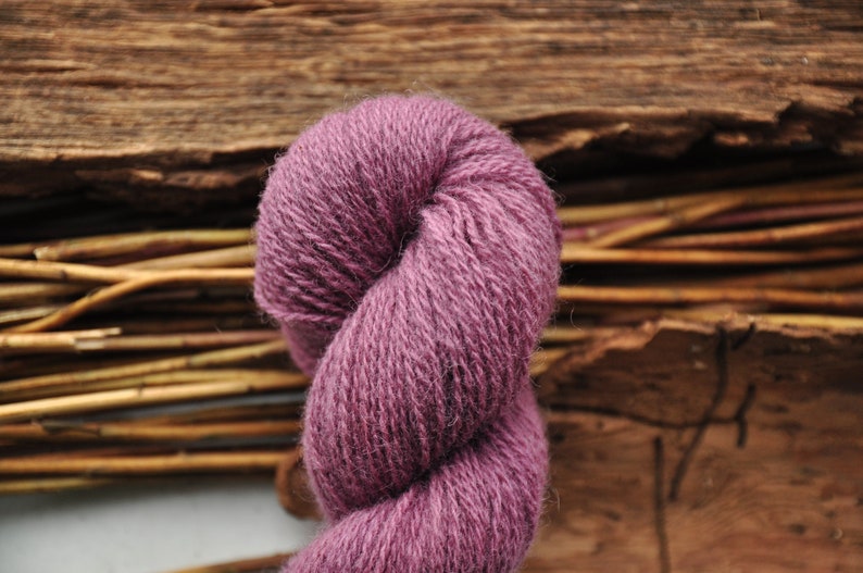 May include: A skein of soft, purple yarn, twisted into a loop. The yarn is made of natural fibers and has a rustic, hand-spun look.