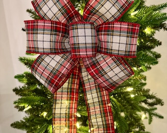 Puede incluir: Un gran lazo decorativo a cuadros rojos, blancos y verdes, sobre un árbol de Navidad. El lazo presenta un patrón de tartán clásico, con largas colas. El árbol está iluminado con pequeñas luces blancas cálidas, creando una escena festiva navideña.