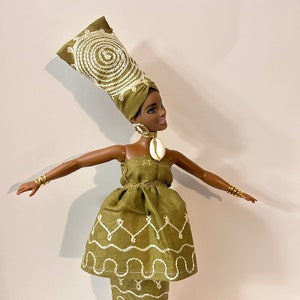 Puede incluir: Mu&ntilde;eca con un vestido verde y un turbante a juego, ambos con detalles bordados en blanco. La mu&ntilde;eca tiene pulseras y pendientes dorados, y sandalias de plataforma blancas. Una concha est&aacute; unida a los pendientes.