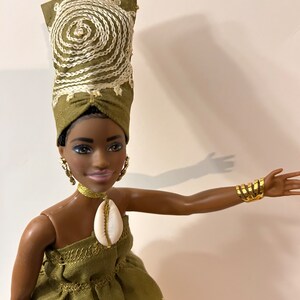 Puede incluir: Mu&ntilde;eca con un vestido verde y un turbante a juego, ambos con bordados blancos. La mu&ntilde;eca tiene pendientes dorados, un collar&iacute;n dorado y un colgante de concha. Una pulsera de pu&ntilde;o dorada adorna el brazo de la mu&ntilde;eca.