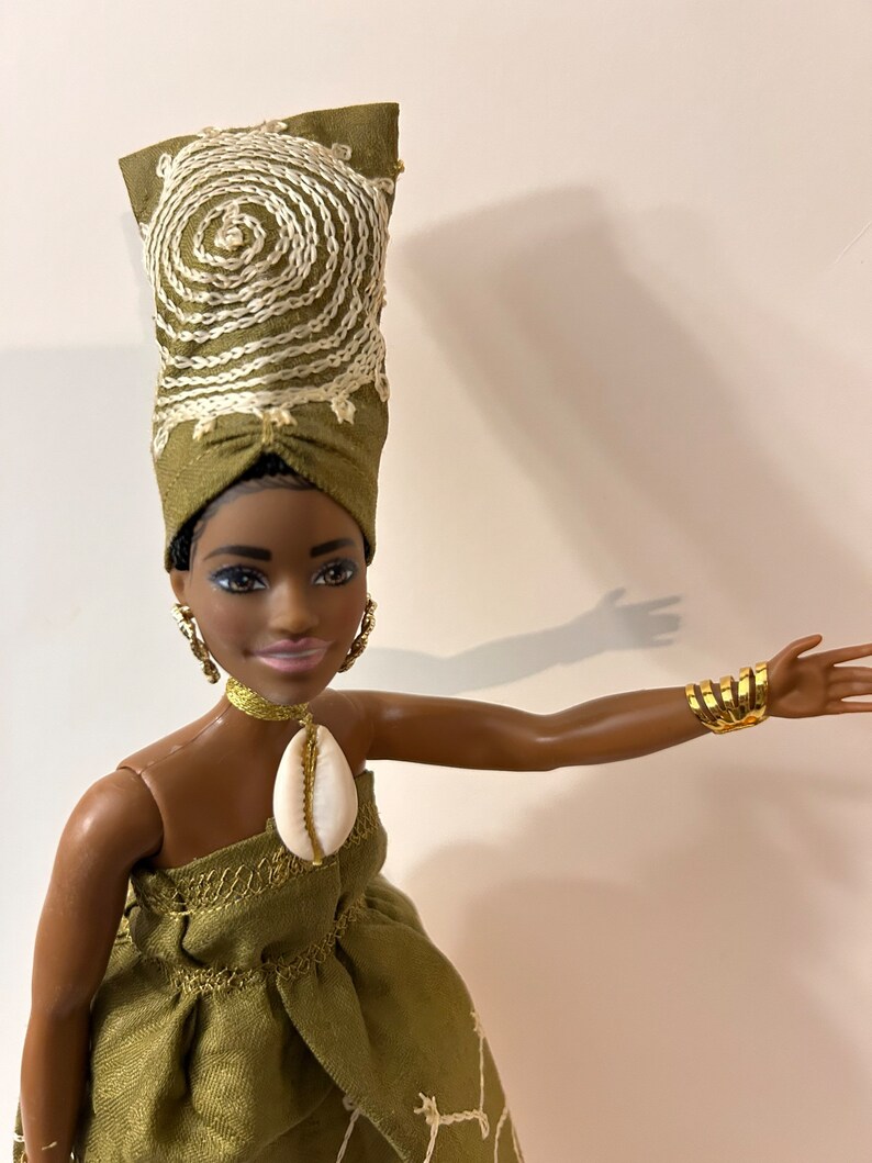 Puede incluir: Mu&ntilde;eca con un vestido verde y un turbante a juego, ambos con bordados blancos. La mu&ntilde;eca tiene pendientes dorados, un collar&iacute;n dorado y un colgante de concha. Una pulsera de pu&ntilde;o dorada adorna el brazo de la mu&ntilde;eca.