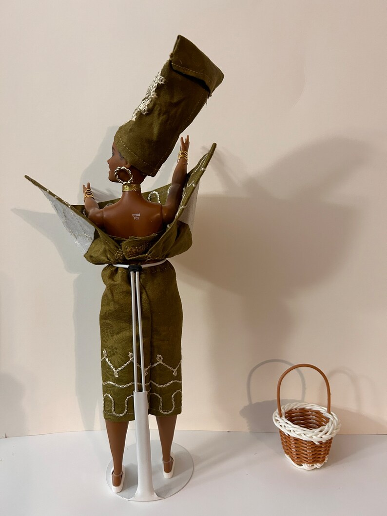 Puede incluir: Mu&ntilde;eca con un atuendo verde oliva con detalles dorados y un tocado a juego. La mu&ntilde;eca est&aacute; sobre un soporte blanco, con una peque&ntilde;a cesta de mimbre cerca. El atuendo tiene un forro plateado y bordados blancos.