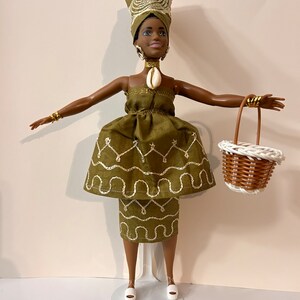 Puede incluir: Mu&ntilde;eca con un vestido verde con detalles bordados blancos, un pa&ntilde;uelo a juego y joyas doradas. La mu&ntilde;eca sostiene una peque&ntilde;a cesta blanca y marr&oacute;n. La mu&ntilde;eca est&aacute; sobre un soporte blanco.