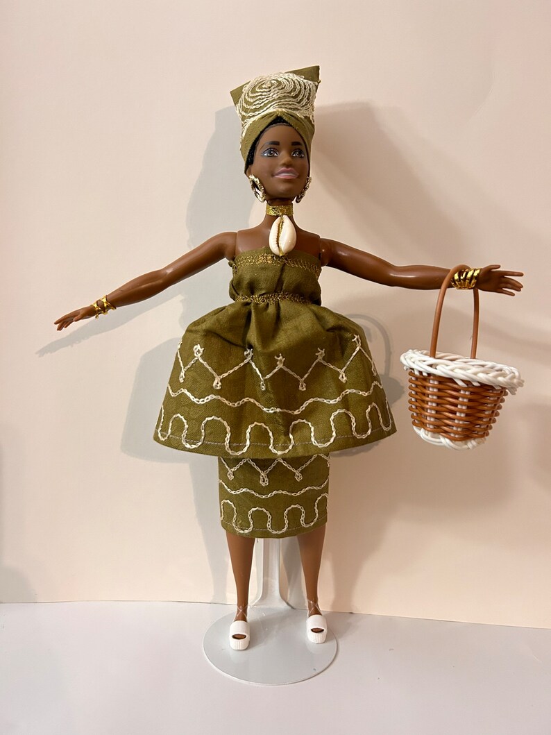 Puede incluir: Mu&ntilde;eca con un vestido verde con detalles bordados blancos, un pa&ntilde;uelo a juego y joyas doradas. La mu&ntilde;eca sostiene una peque&ntilde;a cesta blanca y marr&oacute;n. La mu&ntilde;eca est&aacute; sobre un soporte blanco.