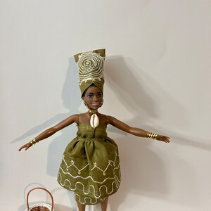 Puede incluir: Mu&ntilde;eca con un vestido verde oliva con bordados blancos, un turbante a juego y pulseras doradas. Una peque&ntilde;a cesta de mimbre est&aacute; al lado. La mu&ntilde;eca tiene un collar de concha y sandalias blancas.