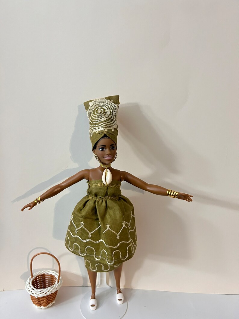 Puede incluir: Mu&ntilde;eca con un vestido verde oliva con bordados blancos, un turbante a juego y pulseras doradas. Una peque&ntilde;a cesta de mimbre est&aacute; al lado. La mu&ntilde;eca tiene un collar de concha y sandalias blancas.