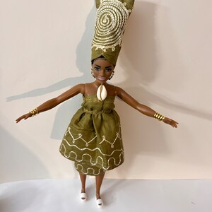 Puede incluir: Mu&ntilde;eca con un vestido verde oliva y un turbante a juego, ambos con detalles bordados en blanco. La mu&ntilde;eca tiene pulseras y pendientes dorados, y un colgante de concha. La mu&ntilde;eca lleva zapatos blancos.