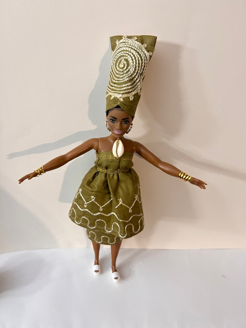 Puede incluir: Mu&ntilde;eca con un vestido verde oliva y un turbante a juego, ambos con detalles bordados en blanco. La mu&ntilde;eca tiene pulseras y pendientes dorados, y un colgante de concha. La mu&ntilde;eca lleva zapatos blancos.
