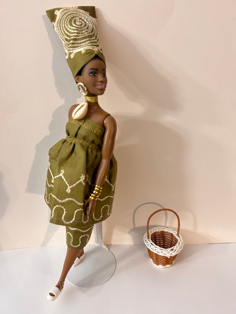 Puede incluir: Una mu&ntilde;eca con un vestido verde oliva con detalles bordados blancos, un turbante a juego y joyas doradas. Una peque&ntilde;a cesta tejida con asa marr&oacute;n y ribete blanco est&aacute; cerca. La mu&ntilde;eca est&aacute; sobre un soporte transparente.