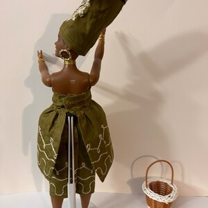Puede incluir: Mu&ntilde;eca con un atuendo verde oliva con joyas doradas y un tocado alto. La mu&ntilde;eca levanta los brazos y tiene una peque&ntilde;a cesta de mimbre al lado. El atuendo tiene detalles bordados en blanco.