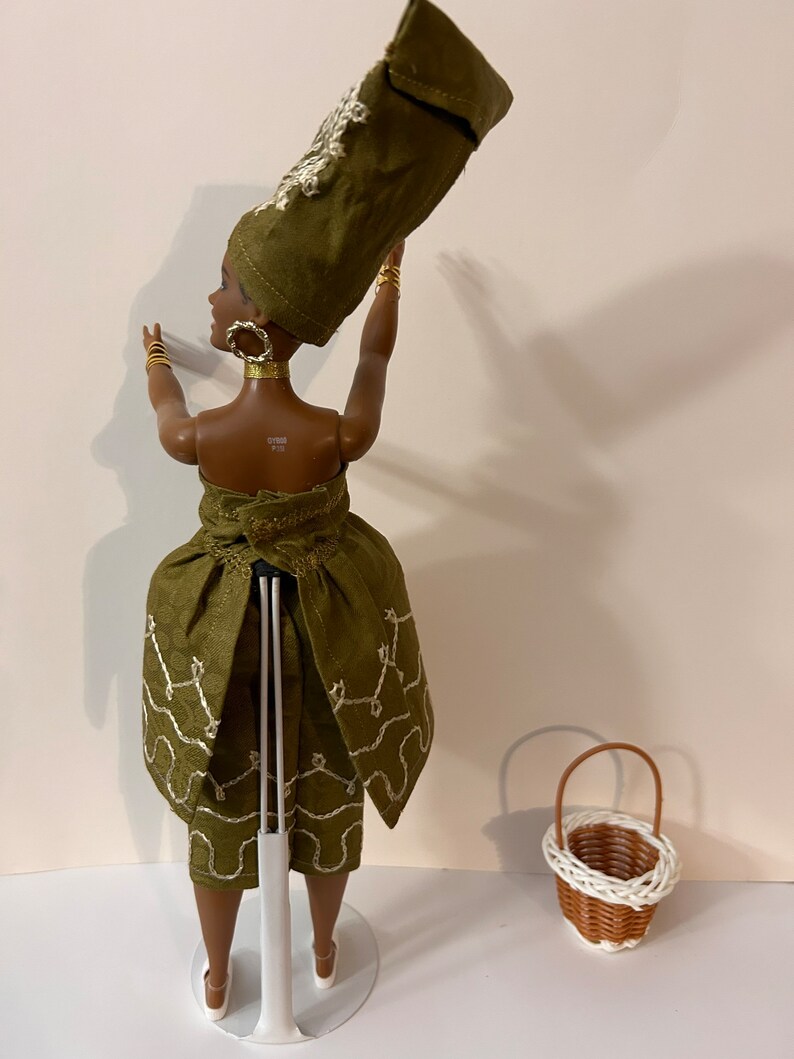 Puede incluir: Mu&ntilde;eca con un atuendo verde oliva con joyas doradas y un tocado alto. La mu&ntilde;eca levanta los brazos y tiene una peque&ntilde;a cesta de mimbre al lado. El atuendo tiene detalles bordados en blanco.