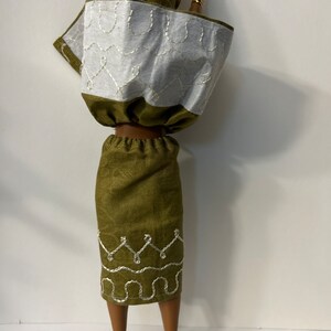 Puede incluir: Una mu&ntilde;eca con un atuendo verde y blanco. El conjunto incluye una falda verde con detalles bordados en blanco, una blusa blanca y verde y un pa&ntilde;uelo a juego. Brazaletes dorados adornan los brazos de la mu&ntilde;eca y zapatos blancos completan el conjunto.