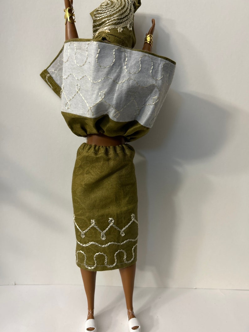 Puede incluir: Una mu&ntilde;eca con un atuendo verde y blanco. El conjunto incluye una falda verde con detalles bordados en blanco, una blusa blanca y verde y un pa&ntilde;uelo a juego. Brazaletes dorados adornan los brazos de la mu&ntilde;eca y zapatos blancos completan el conjunto.