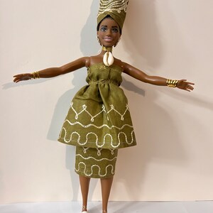 Puede incluir: Mu&ntilde;eca vestida con un vestido verde oliva y un turbante a juego con detalles bordados en blanco. La mu&ntilde;eca tiene un collar de concha, pulseras doradas y zapatos de plataforma blancos. El turbante presenta un dise&ntilde;o en espiral.