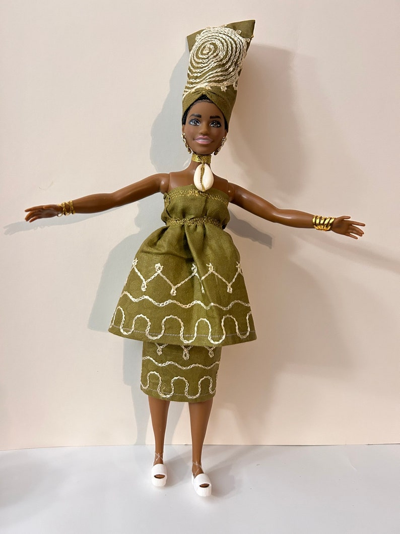 Puede incluir: Mu&ntilde;eca vestida con un vestido verde oliva y un turbante a juego con detalles bordados en blanco. La mu&ntilde;eca tiene un collar de concha, pulseras doradas y zapatos de plataforma blancos. El turbante presenta un dise&ntilde;o en espiral.