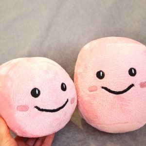 May include: Two pink plush toys shaped like cubes with embroidered smiling faces. The larger toy has a wider smile and blush marks on its cheeks.