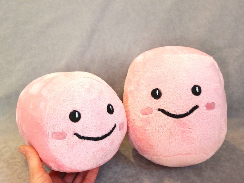 May include: Two pink plush toys shaped like cubes with embroidered smiling faces. The larger toy has a wider smile and blush marks on its cheeks.