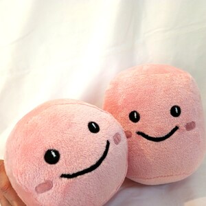 May include: Two pink plush toys with smiling faces. The toys are shaped like a sphere and a cube.