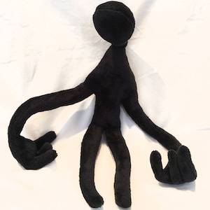 Puede incluir: Juguete de peluche negro con forma de persona con brazos y piernas largos. El juguete est&aacute; sentado sobre una superficie blanca.