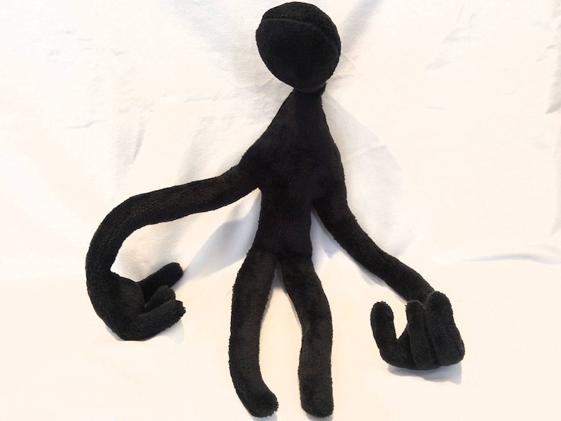 Puede incluir: Juguete de peluche negro con forma de persona con brazos y piernas largos. El juguete est&aacute; sentado sobre una superficie blanca.