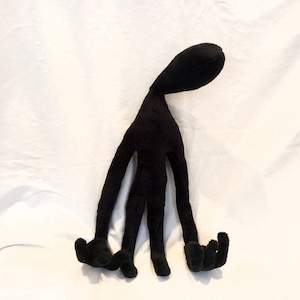 Puede incluir: Juguete de peluche negro con forma de figura humanoide con cuatro brazos largos y un cuello largo. El juguete est&aacute; de pie sobre una superficie blanca.