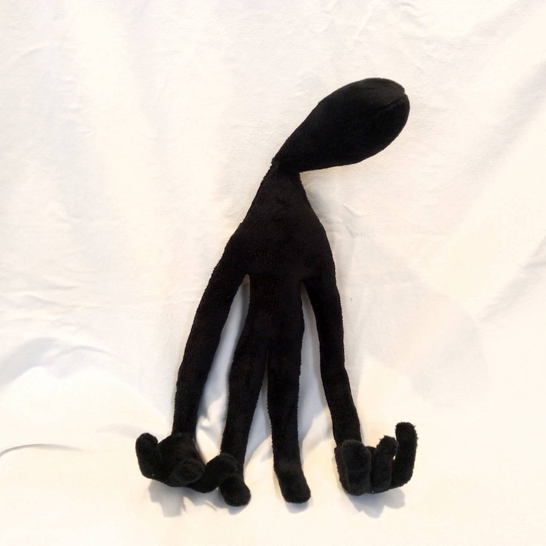 Puede incluir: Juguete de peluche negro con forma de figura humanoide con cuatro brazos largos y un cuello largo. El juguete est&aacute; de pie sobre una superficie blanca.