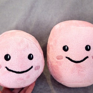 May include: Two pink plush toys with smiling faces. The larger toy is rectangular and the smaller toy is round.
