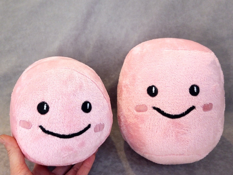 May include: Two pink plush toys with smiling faces. The larger toy is rectangular and the smaller toy is round.
