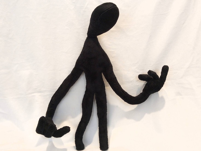 Puede incluir: Juguete de peluche negro con forma de figura humanoide alta y delgada con brazos y piernas largos. El juguete tiene una cabeza grande y redonda y no tiene rasgos faciales.