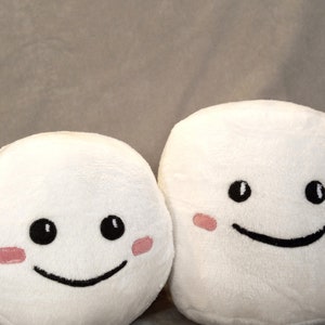 May include: Two white plush toys shaped like marshmallows with black eyes and a black smiling mouth. Each toy has pink blush on its cheeks.