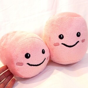 May include: Two pink plush toys shaped like pills with smiling faces. The toys have black eyes and mouths.
