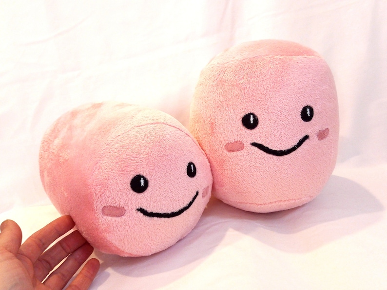 May include: Two pink plush toys shaped like pills with smiling faces. The toys have black eyes and mouths.
