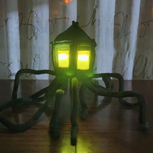 Peut inclure: Une lampe verte en feutre en forme de maison avec des fenêtres jaunes qui brillent et de multiples tentacules. La lampe est posée sur une surface en bois.