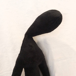 Puede incluir: Juguete de peluche negro con forma de persona, cuello largo y cabeza inclinada hacia abajo. El juguete est&aacute; hecho de tela suave y peluda.