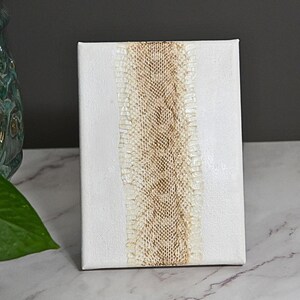 May include: A rectangular white canvas featuring a vertical strip of textured, light brown, snakeskin-patterned material running down the centre. The canvas has a smooth, matte finish and is positioned upright, displaying a clean, neutral aesthetic.