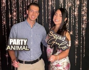 May include: A person in a light blue checkered button-down shirt and khaki pants holding a black sign that says PARTY ANIMAL, and a person in a floral print satin dress holding a black sign that says Squad Goals against a black backdrop.