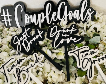 Puede incluir: Letreros de acrílico blanco y negro con varias frases de temática nupcial, incluyendo "#CoupleGoals", "Just Drunk in Love", "Team Bride" y "Team Groom".