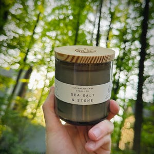 May include: A glass jar candle with a wooden lid is held in front of a blurred green background. The candle has a white label with the text "ALTERNATIVE WAX CANDLE CO SEA SALT & STONE". The candle is olive green.