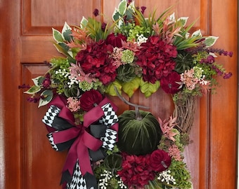 May include: A fall-themed wreath with a grapevine base, adorned with burgundy hydrangeas, green velvet pumpkins, and a large bow with black and white checkered accents. The wreath is decorated with various greenery and small floral elements.