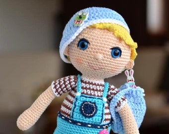 Könnte beinhalten: Eine gehäkelte Puppe trägt ein blau-weiß gestreiftes Hemd, eine blaue Latzhose, einen blauen Hut und blaue Schuhe. Die Puppe hält eine blaue Tasche mit einem weißen Hasen darauf. Die Puppe steht auf einer weißen Oberfläche. "Háčkování a pletení od Yvetty" steht auf der Oberfläche.