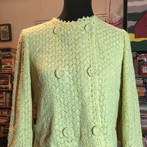 May include: A light yellow, vintage-style jacket with a textured, open-weave design. The jacket features a round collar, long sleeves, and decorative buttons. The jacket is displayed on a black mannequin.