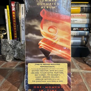 May include: A sealed 1988 Summer Olympics Album compact disc titled One Moment In Time. The cover features a flaming torch graphic and a yellow sticker listing twelve record-breaking performances by artists including Whitney Houston and the Bee Gees.