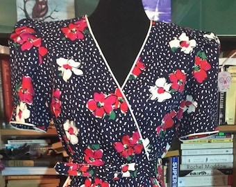 May include: A navy blue wrap dress with a white and pink floral print. The dress has a white trim and a belt. The dress is on a mannequin and is in front of a bookshelf.