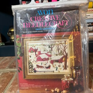 May include: An Avon Creative Needlecraft crewel embroidery kit featuring a winter country scene with red barns and figures. The kit includes a stamped design on 100% cotton, measuring 18" x 22". The package is sealed in plastic.
