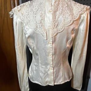 May include: Ivory-colored satin blouse with a high, button-up collar and long sleeves. The blouse features a delicate lace yoke and shoulder detail, with small buttons running down the back. The fabric has a subtle sheen, suggesting a vintage or formal style.