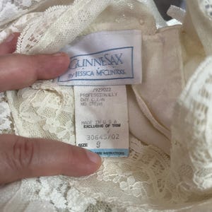 May include: Close-up of a cream-colored lace dress with a tag that reads "GUNNESAX by JESSICA MCCLINTOCK." The tag also indicates the dress is size 9 and made in the USA. The dress has intricate lace detailing.
