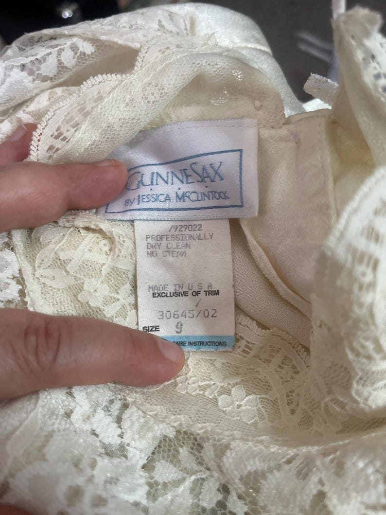 May include: Close-up of a cream-colored lace dress with a tag that reads "GUNNESAX by JESSICA MCCLINTOCK." The tag also indicates the dress is size 9 and made in the USA. The dress has intricate lace detailing.