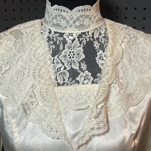 May include: Ivory-colored vintage dress with a high, lace-trimmed collar and a delicate lace overlay. The dress features a satin-like fabric, creating a smooth, elegant appearance. The lace detailing adds a touch of vintage charm to the garment.