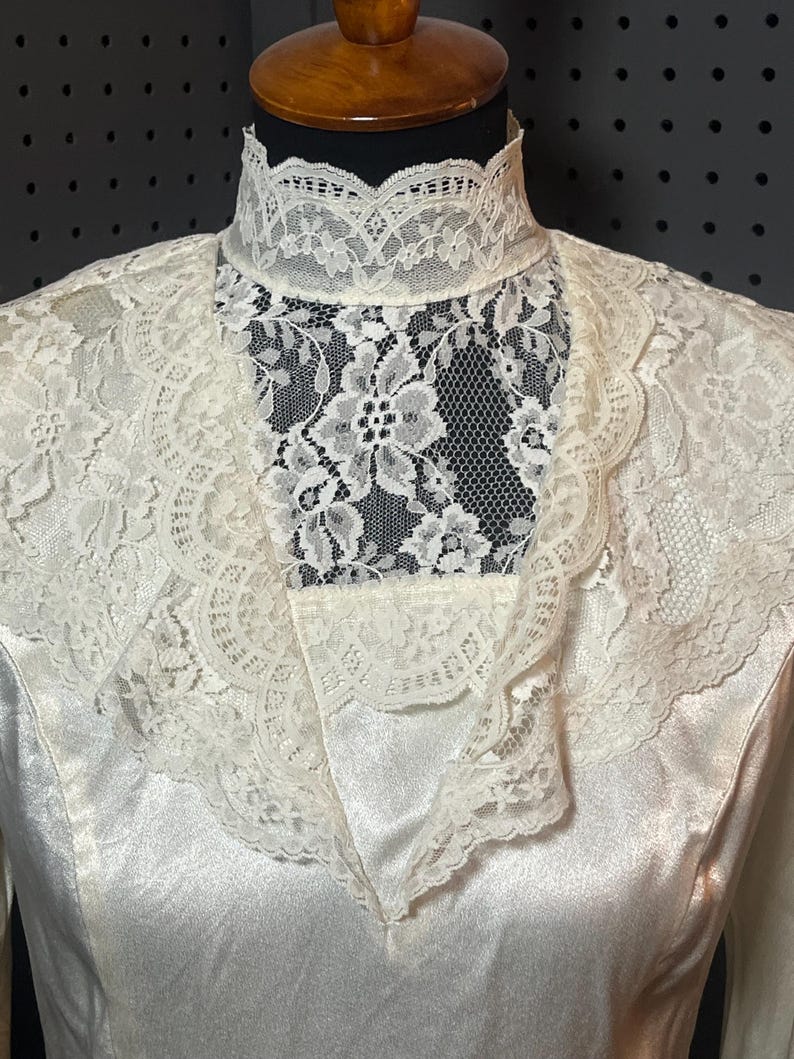 May include: Ivory-colored vintage dress with a high, lace-trimmed collar and a delicate lace overlay. The dress features a satin-like fabric, creating a smooth, elegant appearance. The lace detailing adds a touch of vintage charm to the garment.