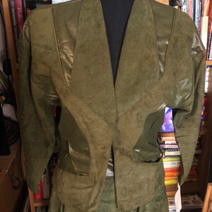May include: A green suede jacket and skirt set with a faux leather trim. The jacket has a V-neck and long sleeves. The skirt is a-line and has a pleated design.
